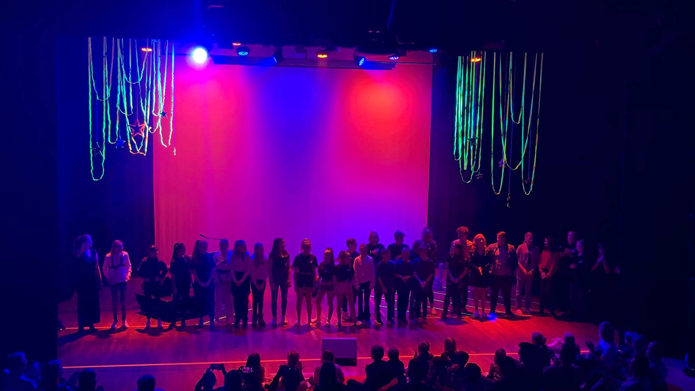 Bilde av en scene med en lang rekke barn og unge som står på rekke i farget scenelys. Publikum applauderer med ryggen til kameraet.