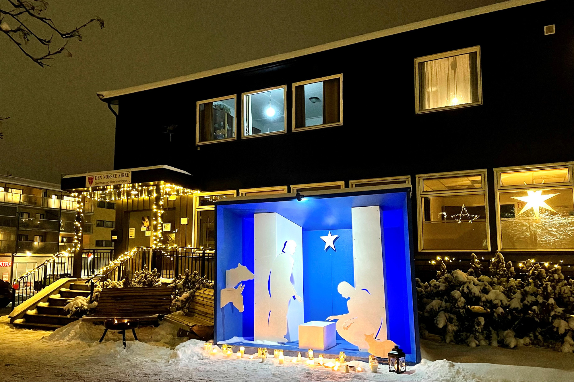 Bilde av en opplyst utendørs julekrybbe. Det er natt og snø på bakken. Krybben er et opplyst rom med blå vegger, med hvite utskårede figurer i full størrelse av en mann og en kvinne som bøyer seg over en hvit kloss som symboliserer krybben. Krybben står på fortauet utenfor et hus med julelys og det er plassert grupper av gravlys og lykter i snøen.